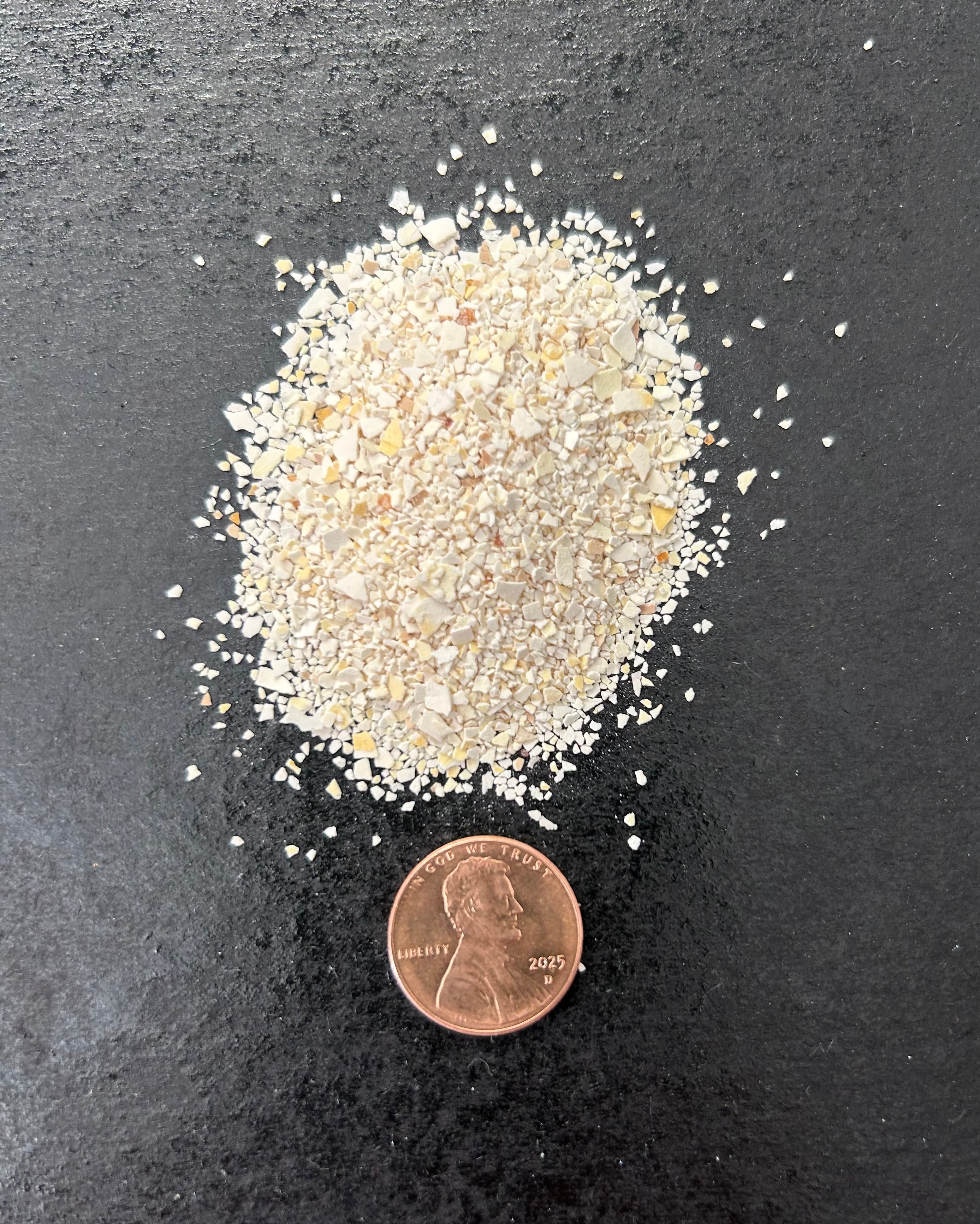 Small pile of ground eggshells next to a penny on a dark surface